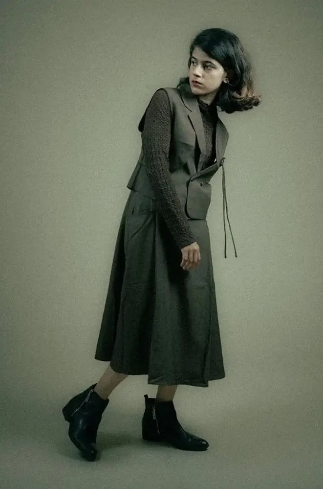 Woman in an olive-grey belted coat and ankle boots against a muted green wall