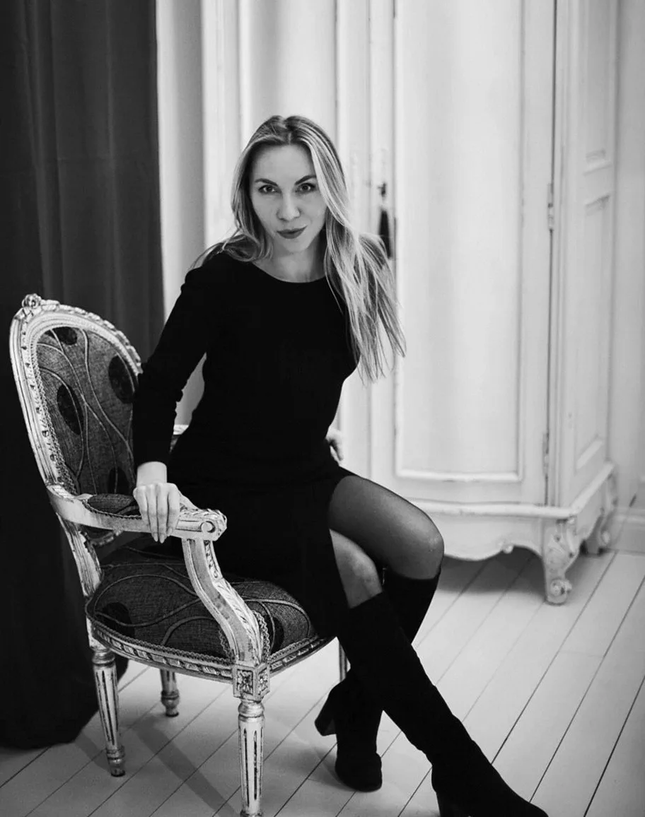 Black-and-white photo of a woman in a black dress and knee-high boots seated on an ornate chair