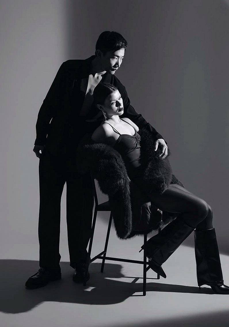 Black-and-white studio photo of two people in dark clothes, the woman seated in a fur-trimmed coat