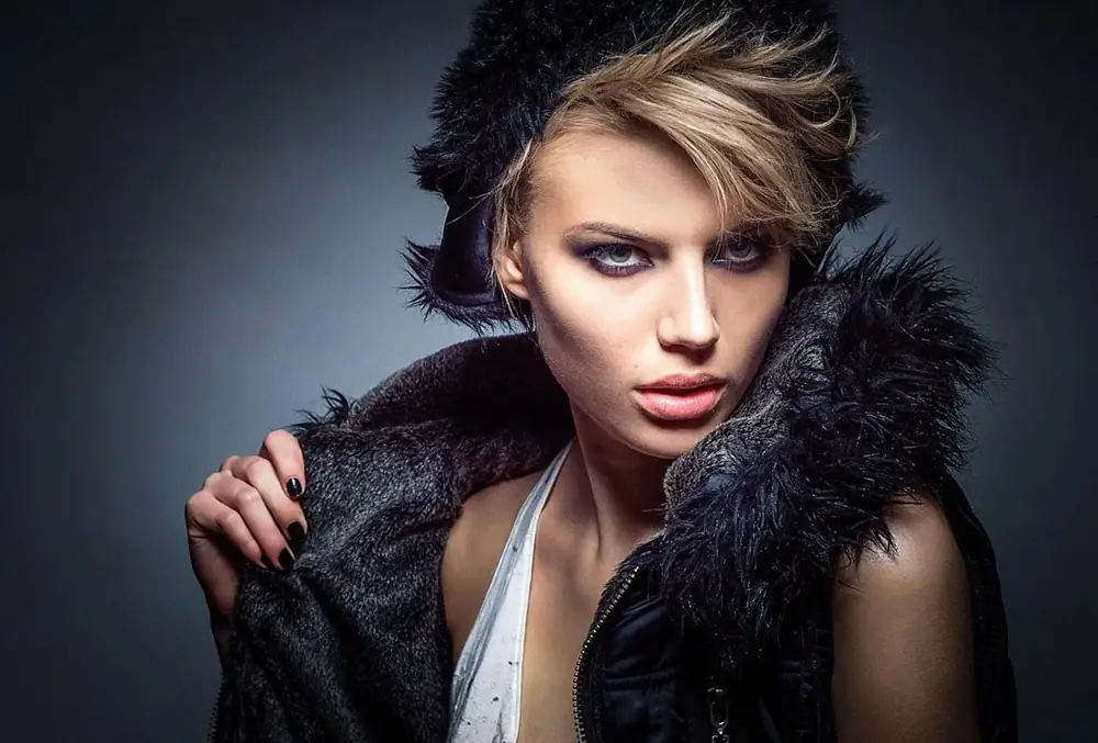 Woman with short wavy hair wearing a black coat with a feather-trimmed collar